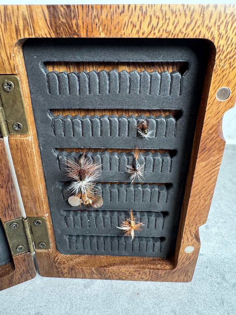 K&ouml;nnte beinhalten: Eine h&ouml;lzerne Fliegenfischbox mit einem schwarzen Schaumstoffeinsatz. Die Box ist ge&ouml;ffnet und es befinden sich mehrere Fliegen im Schaumstoff.