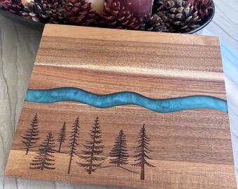 May include: A rectangular wooden serving board featuring a horizontal wavy inlay of translucent turquoise resin. The lower section of the board is decorated with a laser-engraved forest scene consisting of several pine trees of varying heights.