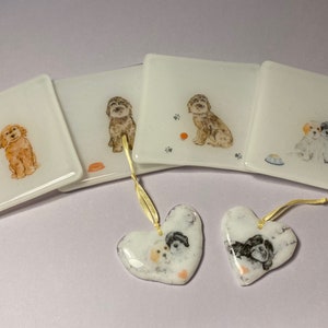 May include: Four white square glass coasters with dog illustrations and two white heart-shaped glass ornaments with dog illustrations. The coasters have gold string attached to them.