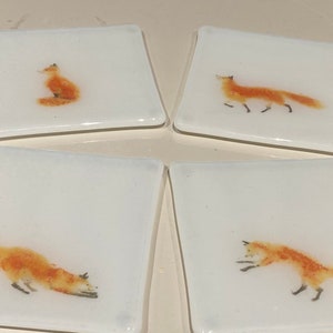 Könnte beinhalten: Vier quadratische Untersetzer aus weißer Keramik mit einem Fuchs-Design. Jeder Untersetzer zeigt eine andere Fuchs-Pose, darunter ein sitzender Fuchs, ein laufender Fuchs und zwei springende Füchse.