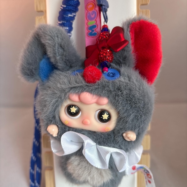 May include: A plush keychain featuring a doll in a gray bunny costume with red and blue accents. The doll has large eyes and a white bow. A pink tag with the word "MIGO" is attached to the keychain.