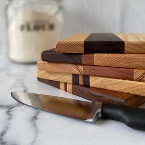 Stake of 5 wooden cutting board ends showing the different types of wood on a white marble countertop. In front of them is a knife. Behind it is a canister of flour. There is a shiplap wooden background.