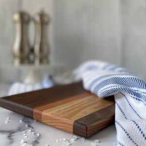 Striped wooden cutting board on a white marble countertop with sea salt scattered in front of board. There is a blue and white striped towel around the right of the board. Plus silver salt and pepper skaters and white shiplap in the background.