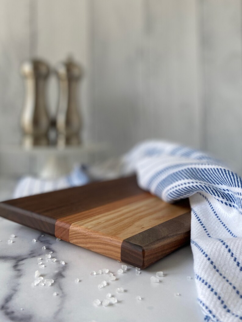 Striped wooden cutting board on a white marble countertop with sea salt scattered in front of board. There is a blue and white striped towel around the right of the board. Plus silver salt and pepper skaters and white shiplap in the background.
