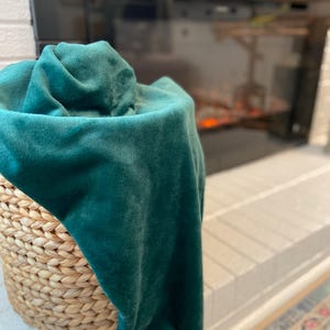 May include: A woven wicker basket holds a folded, emerald green velvet throw blanket. The blanket's plush texture contrasts with the basket's natural, textured surface. The basket sits on a white brick ledge.