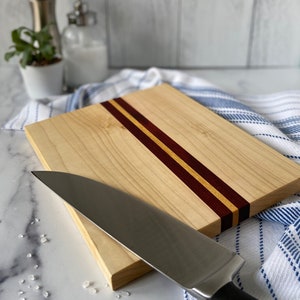 Center striped wooden cutting board on a white marble countertop with sea salt scattered in front of board and a knife resting on the board. There is a blue & white striped towel under the board. There is a white ship-lapped background.