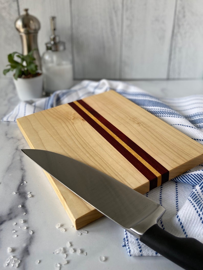 Center striped wooden cutting board on a white marble countertop with sea salt scattered in front of board and a knife resting on the board. There is a blue & white striped towel under the board. There is a white ship-lapped background.