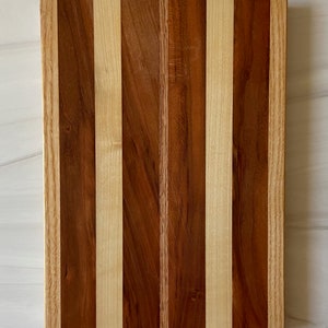 Puede incluir: Una tabla de cortar rectangular de madera con un patr&oacute;n de rayas de madera clara y oscura. La tabla est&aacute; hecha de varios tipos de madera, incluyendo arce y nogal. La tabla est&aacute; etiquetada con la letra "F".