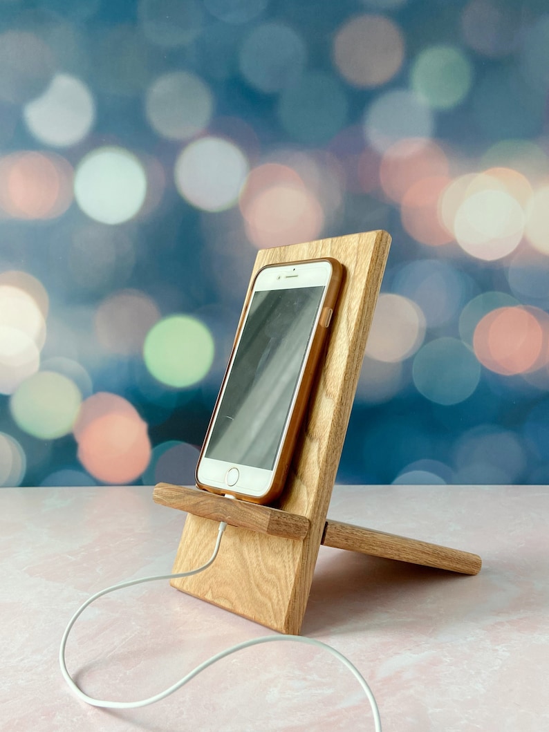 A wooden cell phone stand showing how you can hold your cell phone and charge it at the same time, on a pink marble table with pink and blue pastel background