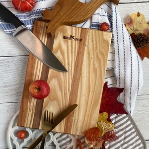 Striped wooden cutting board on a ship-lapped base. It is surrounded by a blue and white striped towel, knife, gold utensils, pot holders, oven safety deck, fall leaves, small pumpkins, and an apple.