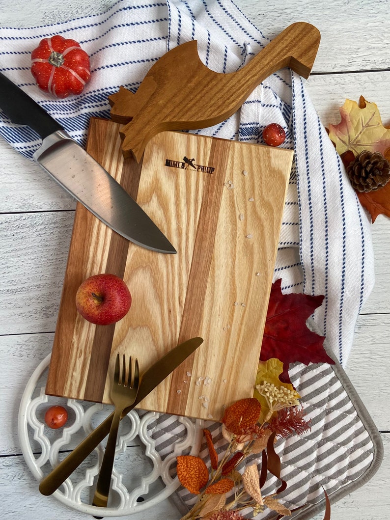 Striped wooden cutting board on a ship-lapped base. It is surrounded by a blue and white striped towel, knife, gold utensils, pot holders, oven safety deck, fall leaves, small pumpkins, and an apple.