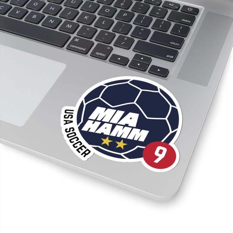 May include: A sticker featuring a soccer ball design with the text "MIA HAMM" and "USA SOCCER." The sticker includes two gold stars and the number 9 in a red circle. The soccer ball is navy blue.