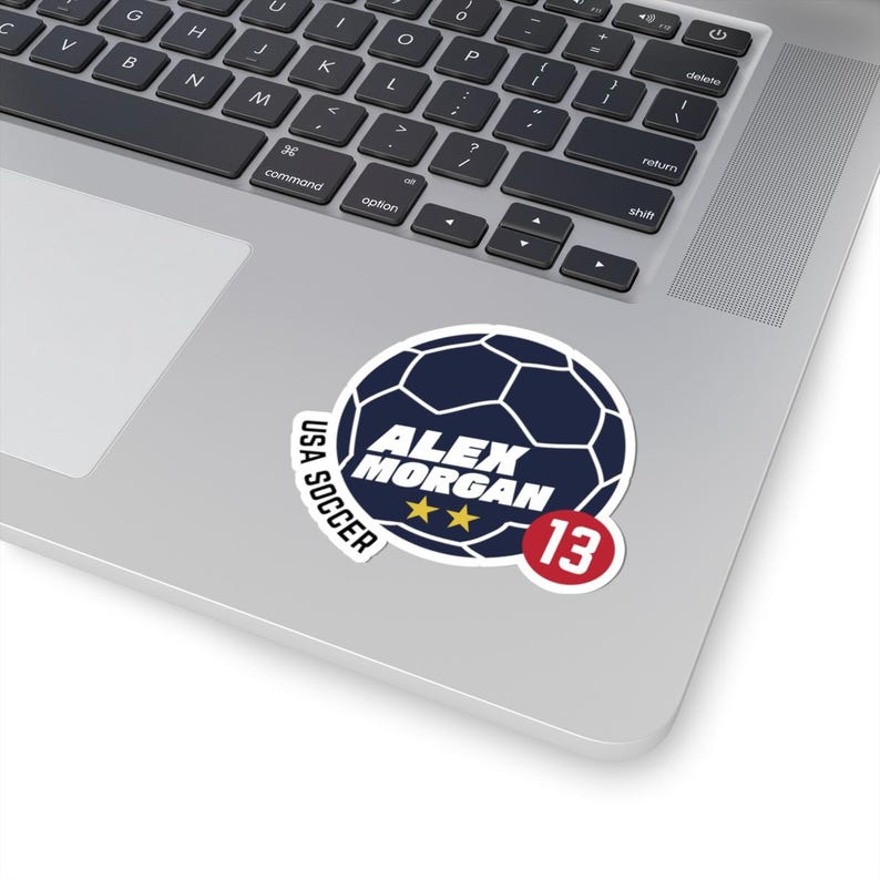 May include: A white sticker featuring a navy blue soccer ball design with the text "ALEX MORGAN" and two gold stars. Below the name is the number "13" in a red circle. The words "USA SOCCER" are also included.