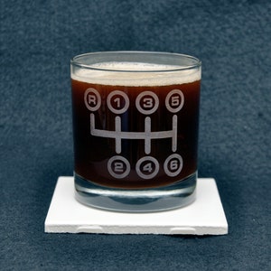 Puede incluir: Un vaso de vidrio transparente con un líquido marrón en su interior. El vaso tiene un diseño blanco de un patrón de cambio de marchas de un coche con las letras R, 1, 2, 3, 4, 5 y 6.