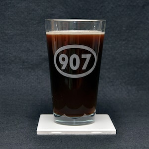 May include: A clear glass pint glass with a dark brown liquid inside. The glass has a white oval etched on the side with the number "907" inside.