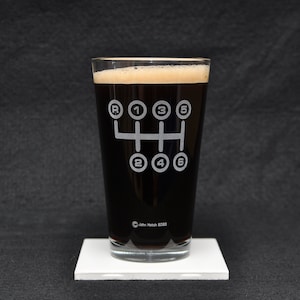 Puede incluir: Un vaso de pinta de vidrio transparente con un líquido negro en su interior. El vaso tiene un diseño grabado en blanco de un patrón de cambio de marchas de transmisión manual de 6 velocidades.