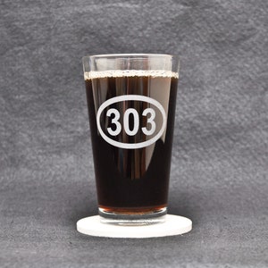 May include: A clear glass pint glass with a black beverage inside. The glass has a white oval etched on the side with the number "303" inside.