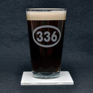 May include: A clear glass pint glass filled with a dark brown liquid. The glass has a white oval etched on the side with the number "336" inside.