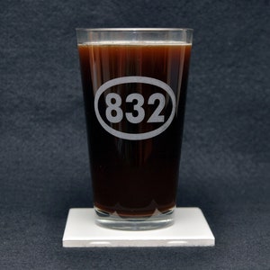 May include: A clear glass pint glass with a dark brown liquid inside. The glass has a white oval etched on the side with the number "832" inside.