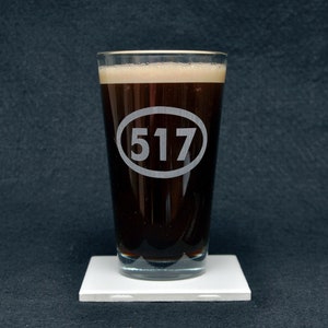 May include: A clear glass pint glass with a dark brown liquid inside. The glass has a white oval etched on the side with the number "517" inside.