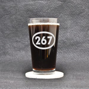 Könnte beinhalten: Ein klares Glas Pint-Glas mit einer dunklen Flüssigkeit darin. Das Glas hat einen weißen Kreis mit der Nummer "267" darauf gedruckt. Das Glas steht auf einem weißen Untersetzer.