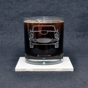 May include: A clear glass tumbler with a dark liquid inside. The glass has an etched design of a classic car on the front. The text "John Katch 2020" is etched on the bottom of the glass.