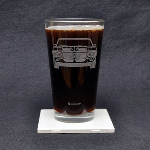 May include: A clear glass pint glass with a dark brown liquid inside. The glass has an etched design of a classic muscle car on the front.