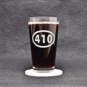 May include: A clear glass pint glass with a black beverage inside. The glass has a white oval with the number "410" printed on it. The glass is sitting on a white coaster.
