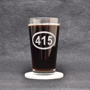 Op de afbeelding: Een helder glazen pintglas met een zwarte vloeistof erin. Het glas heeft een witte ovaal met het nummer "415" erop gedrukt. Het glas staat op een witte onderzetter.