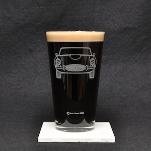 May include: A clear glass pint glass with a dark brown liquid inside. The glass has a white outline of a classic car etched into the side. The text "John Hetch 8080" is printed on the bottom of the glass.