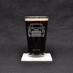 Peut inclure: Un verre de type pinte rempli d'une boisson sombre avec de la mousse, orné d'un dessin blanc gravé d'une voiture. Le verre est posé sur un dessous de verre carré blanc. Le texte "John Hatch 2020" est visible.