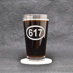 May include: A pint glass filled with dark liquid, possibly beer, resting on a white coaster. The glass has the number "617" in a white oval. The background is a dark gray fabric.