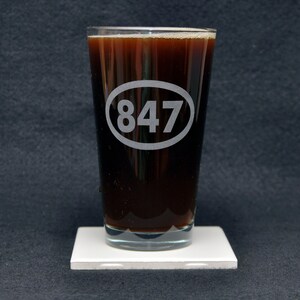 May include: A clear glass pint glass with a dark brown liquid inside. The glass has a white oval etched on the side with the number "847" inside.