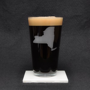 Puede incluir: Un vaso de pinta con cerveza oscura y un contorno blanco del estado de Nueva York grabado en el lateral. El vaso está sobre un posavasos blanco.