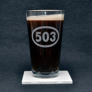 Op de afbeelding: Een helder glazen pintglas met een donkerbruine vloeistof erin. Het glas heeft een witte ovale gravure aan de voorkant met het nummer "503" erin.