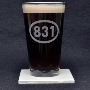 Puede incluir: Un vaso de pinta de vidrio transparente con un líquido marrón oscuro en su interior. El vaso tiene un óvalo blanco grabado en el lateral con el número "831" en su interior.