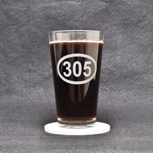 Puede incluir: Un vaso de vidrio transparente con un líquido oscuro en su interior. El vaso tiene un óvalo blanco con el número "305" impreso en él. El vaso está sobre un posavasos blanco.