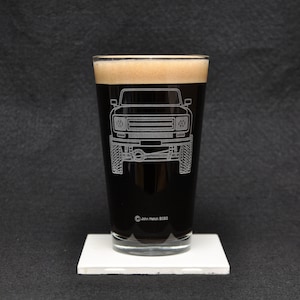 May include: A clear glass pint glass with a black liquid inside. The glass has a white outline of a vintage off-road vehicle etched into the side. The text "John Hatch 2020" is at the bottom of the glass.