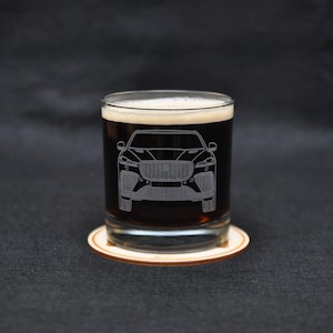 May include: A clear glass with a dark brown liquid inside. The glass has an etched design of a car on the front. The glass is sitting on a wooden coaster.