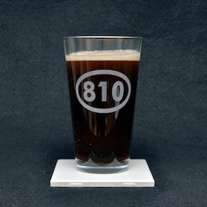 May include: A glass of dark beer in a pint glass with the number 810 etched on the side. The glass is sitting on a white coaster.