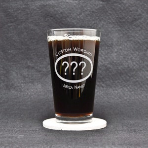 Könnte beinhalten: Ein klares Pintglas, gefüllt mit einer dunklen Flüssigkeit, möglicherweise Bier oder Stout, steht auf einem weißen Untersetzer. Das Glas hat einen geätzten Text: "CUSTOM WORDING" über einem Oval mit drei Fragezeichen und "-AREA NAME-" darunter.
