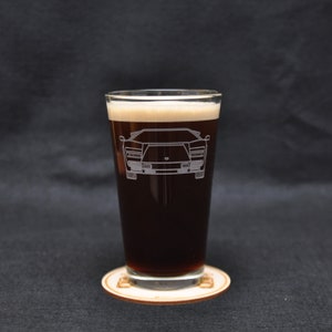 May include: A clear glass pint glass with a dark brown liquid inside. The glass has an etched design of a white Lamborghini Countach.