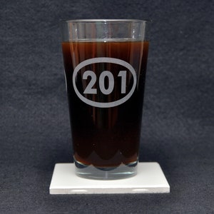 以下が含まれることがあります： 透明なガラス製のピントグラスに、濃い茶色の液体が注がれています。グラスの側面には、白い楕円形がエッチングされており、その中に「201」という数字が刻まれています。