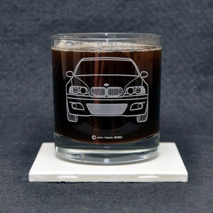 May include: A clear glass tumbler with a dark liquid inside. The glass has a white outline of a BMW M3 car etched into the side. The glass is sitting on a white coaster.