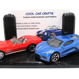 May include: A black business card holder with three toy cars: a red convertible, a blue sports car, and a blue convertible. The holder has a white card with the text "COOL CAR CRAFTS" and contact information.