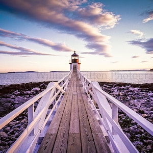 Könnte beinhalten: Ein weißer Holzsteg führt zu einem weißen Leuchtturm auf einer kleinen Insel. Der Himmel ist in wunderschönen Blau- und Rosatönen mit flauschigen Wolken. Das Wasser ist ruhig und blau.