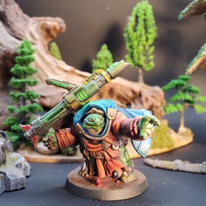 May include: A green and brown miniature figure of a turtle-like creature wearing a blue and orange robe, holding a green and gold weapon. The figure is standing on a round base.