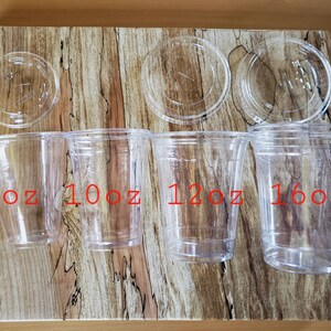 Puede incluir: Cuatro vasos de pl&aacute;stico transparente con tapas, etiquetados 9oz, 10oz, 12oz y 16oz. Los vasos est&aacute;n dispuestos en orden de tama&ntilde;o de izquierda a derecha sobre una superficie de madera.