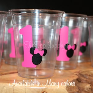 Puede incluir: Vasos de pl&aacute;stico transparentes con pegatinas de Minnie Mouse en rosa y negro. Las pegatinas muestran el n&uacute;mero uno y una cabeza de Minnie Mouse con un lazo rosa. El texto "Disponible en muchos colores" est&aacute; impreso debajo de los vasos.