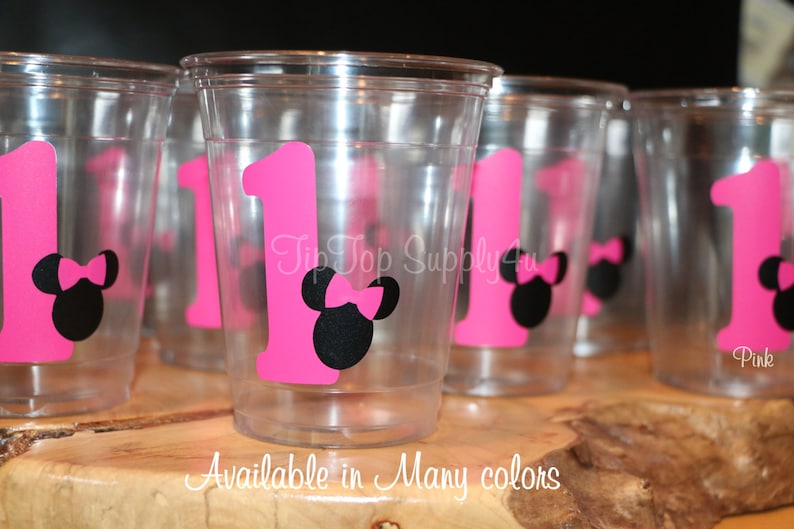 Puede incluir: Vasos de pl&aacute;stico transparentes con pegatinas de Minnie Mouse en rosa y negro. Las pegatinas muestran el n&uacute;mero uno y una cabeza de Minnie Mouse con un lazo rosa. El texto "Disponible en muchos colores" est&aacute; impreso debajo de los vasos.
